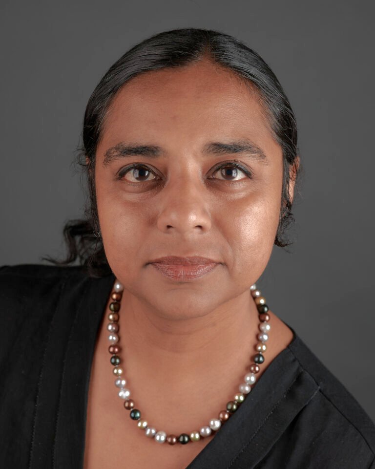 Headshot image of Anna-Liza Badaloo, founder of Anemochory Consulting. She is looking just above the camera lens with a small smile. She is wearing a black top and a necklace of white, light pink and brown pearls.
