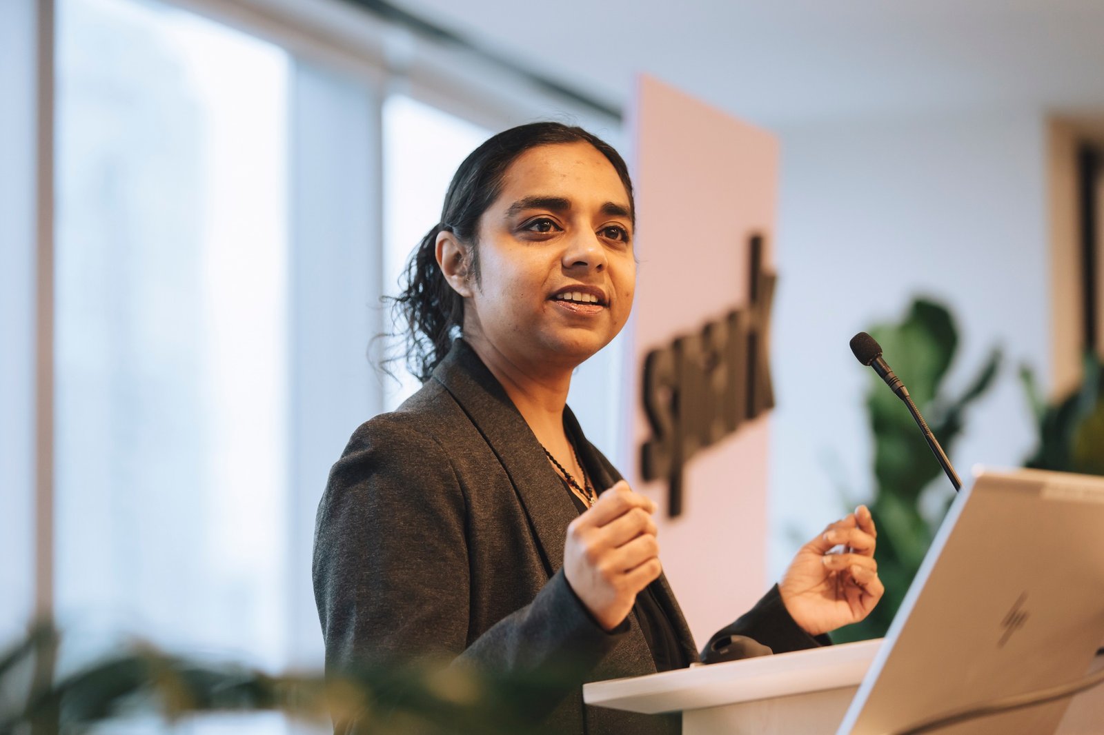 Anna-Liza is standing at a podium speaking at a SPARK Canada event. Her hair is tied back and she is wearing a dark coloured blazer style jacket. A 'spark' banner is behind her out of focus.